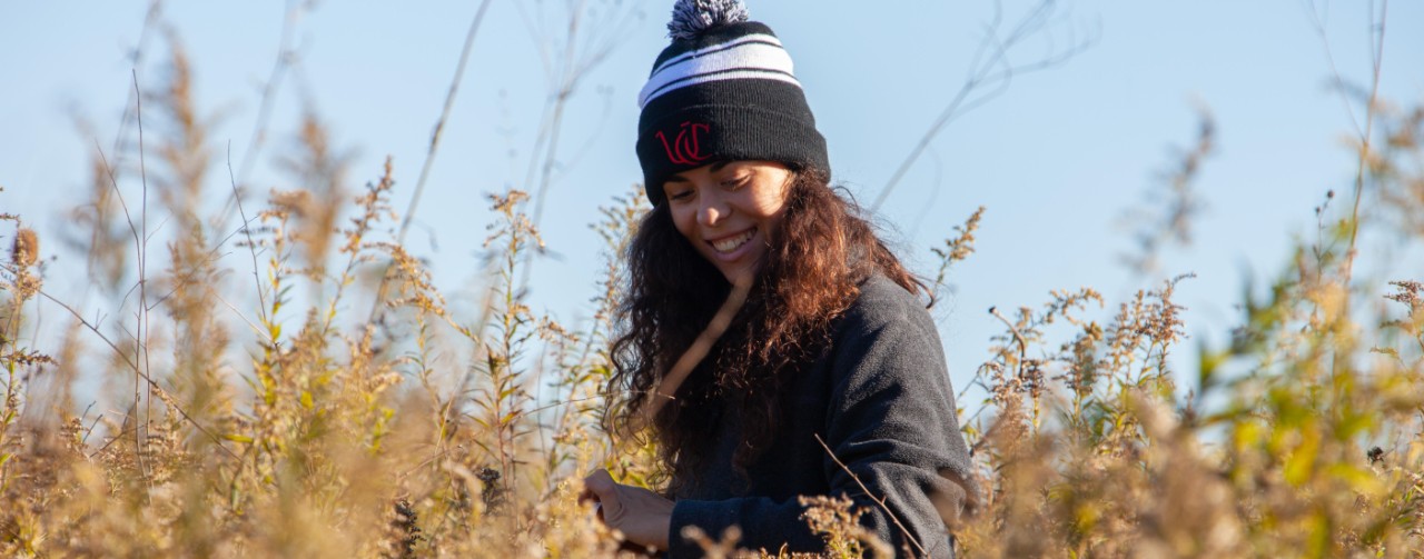 A UC botany student wearing a UC knit cap smiles in a field of yellow goldenrod.