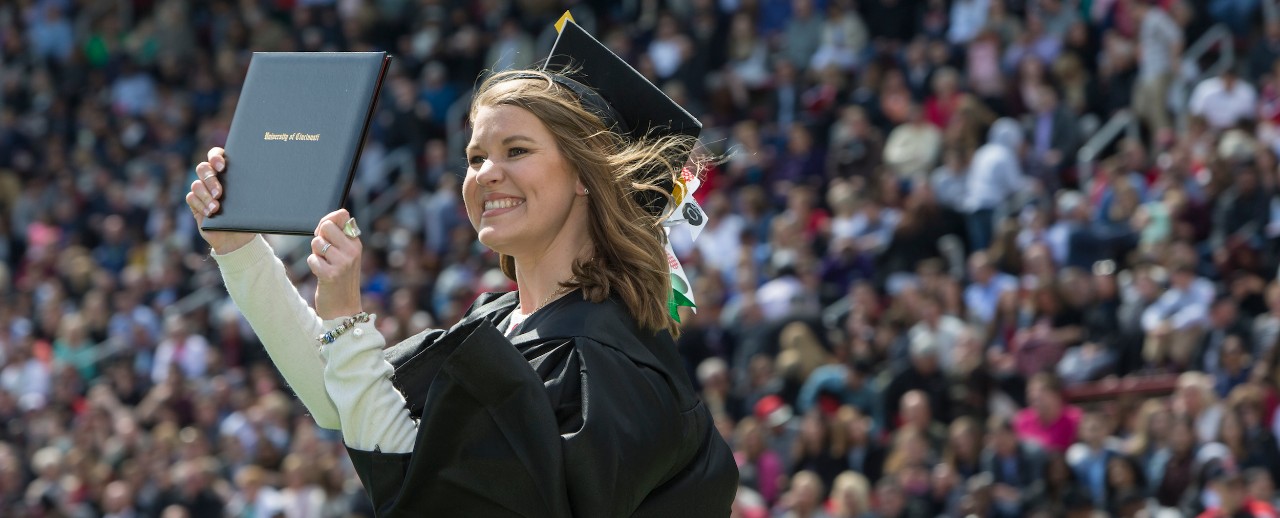 A jubilant UC graduate in a cap and gown waves her diploma for supporters to see as she leaves the stage.