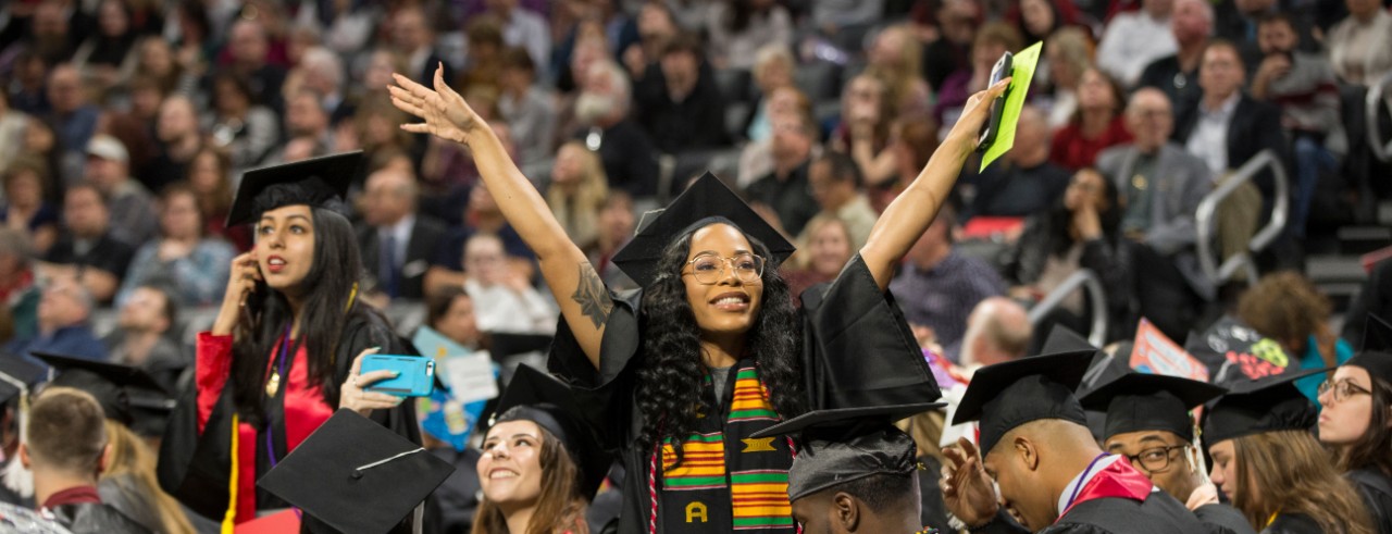 A UC graduate holds her arms up in triumph with seated guests behind her at Fifth Third Arena.