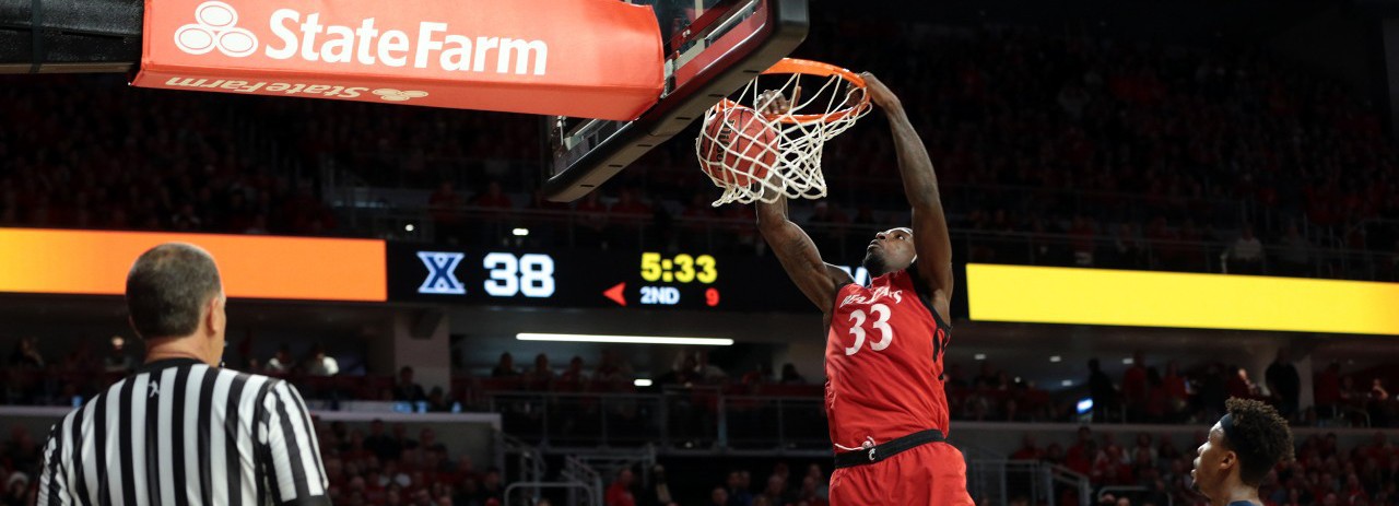 A UC men's basketball player dunks the ball during a game at Fifth Third Arena. A referee in a striped shirt and a Xavier player are also in the photo. 