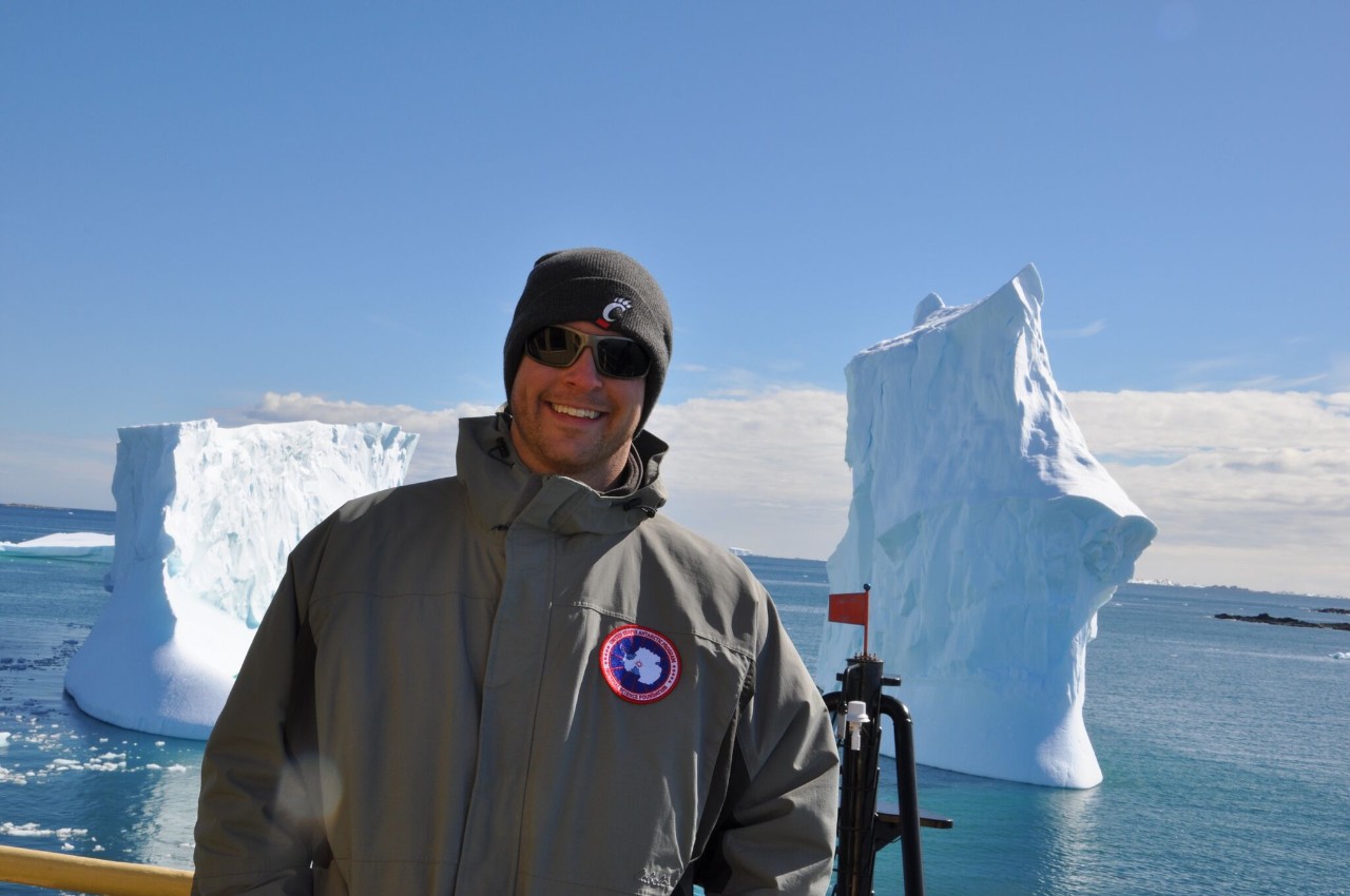 UC professor Joshua Benoit stands on a boat deck in Antarctic with icebergs behind him.