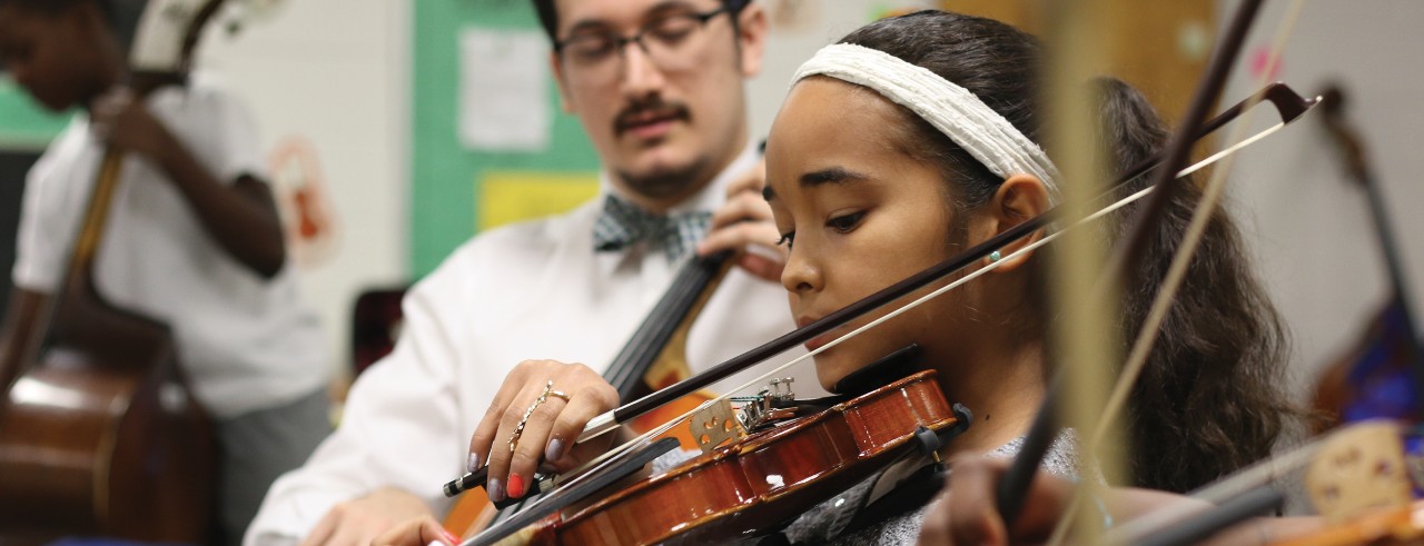 Young students learn how to play string instruments in a classroom.