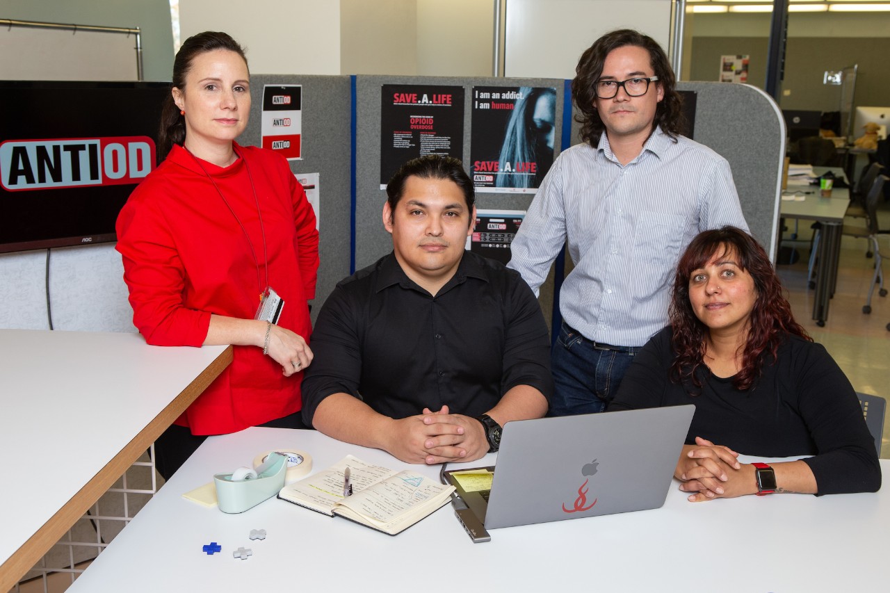 Four people pose in a design lab around a  computer and desk