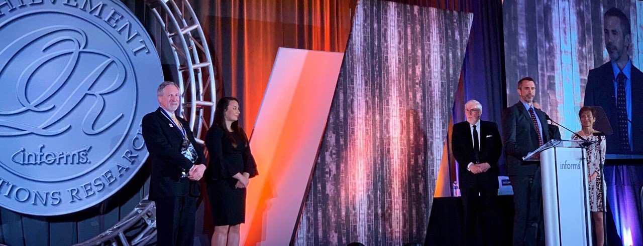 Three men and two woman stand on a backlit stage at an event