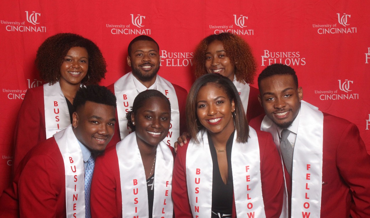 Three young adults stand and smile behind four young adults wearing red blazers and white stoles that say "Business Fellows"