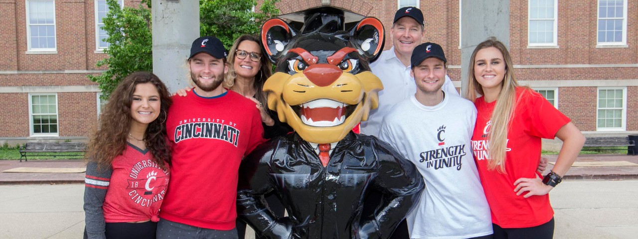 A family of six gathers around a statue of the UC Bearcat dressed in graduation clothing outside of McMicken Hall. 
