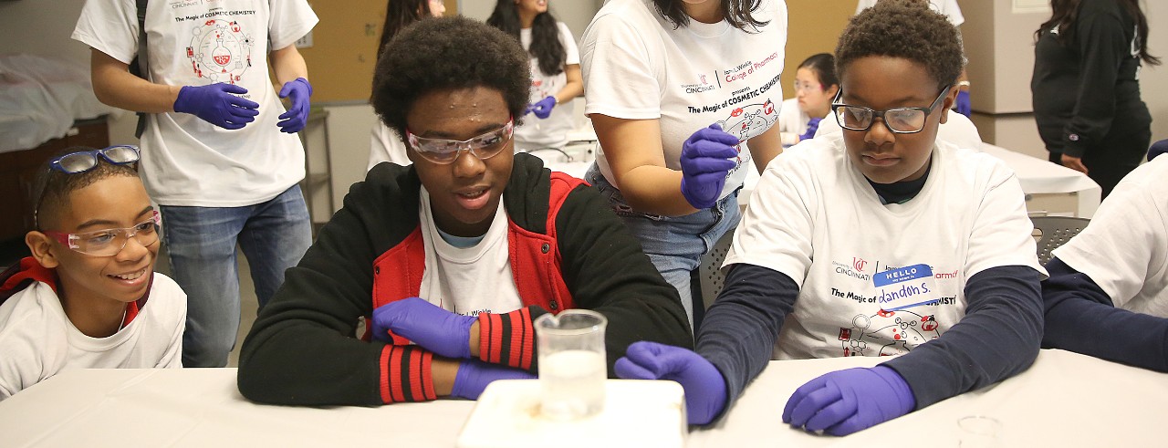Three young high school students are in a lab, wearing goggles and watching as a chemical compound fizzes in a glass beaker. 