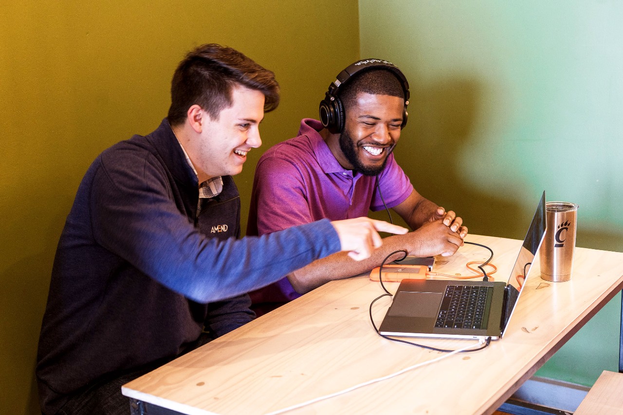 A young man in a navy blue sweater sits next to a young man in a purple collared shirt wearing headphones and look at the same laptop screen
