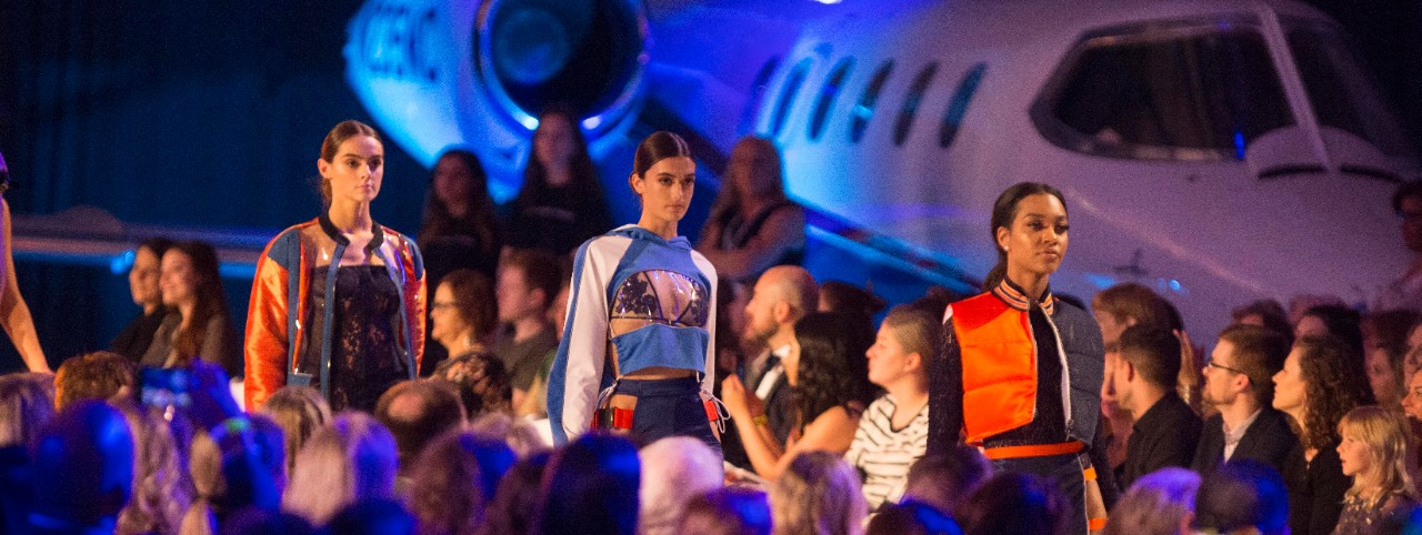 Models walk on a runway in front of a crow with an airplane in the background