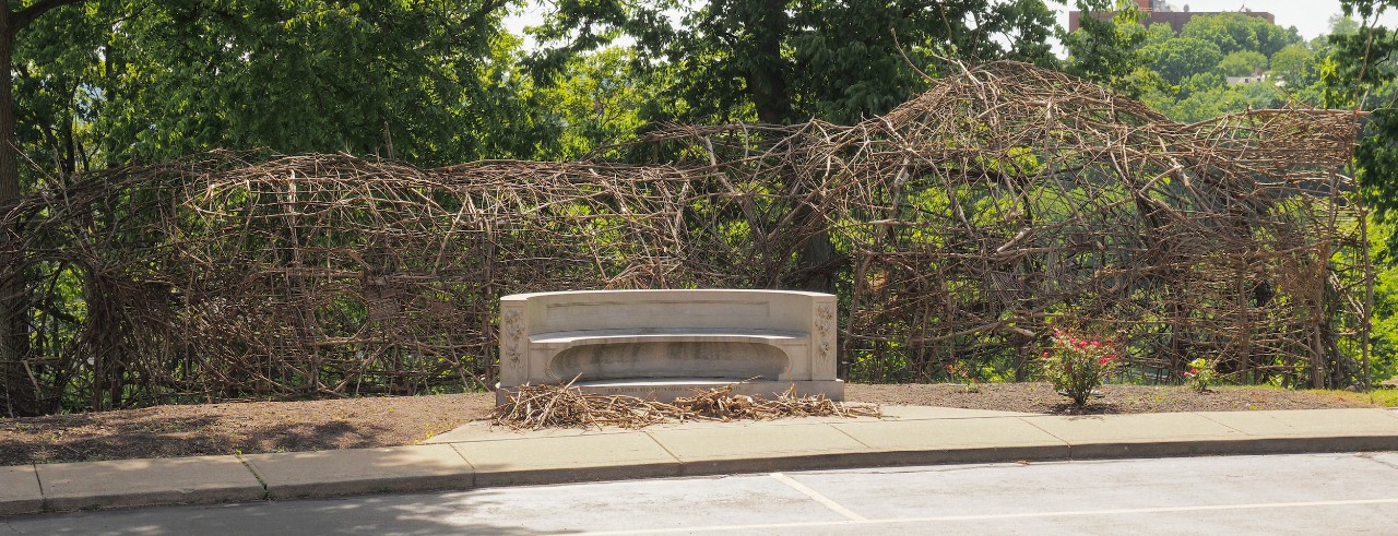 A tunnel-like structure of sticks stands behind a concrete bench outside