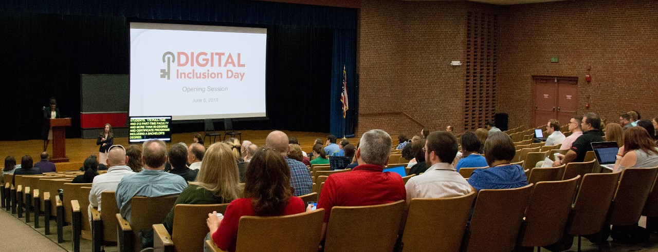 Wide shot of an auditorium with a large audience; a speaker speaks next to a CART writer along with a screen with the title "Digital Inclusion Day"