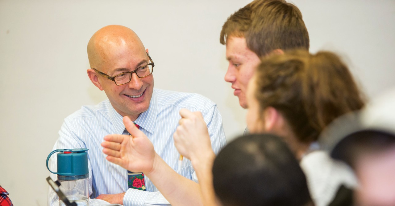 Man wearing glasses sits and speaks to a male student who is motioning with his hands