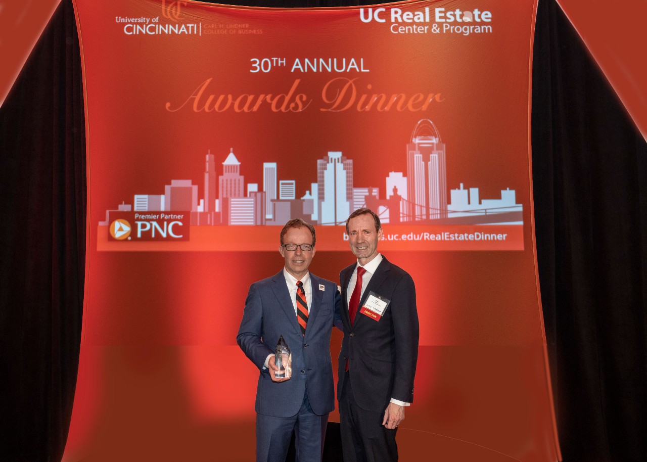 Two men in suits and red ties stand and smile in front of background for real estate center