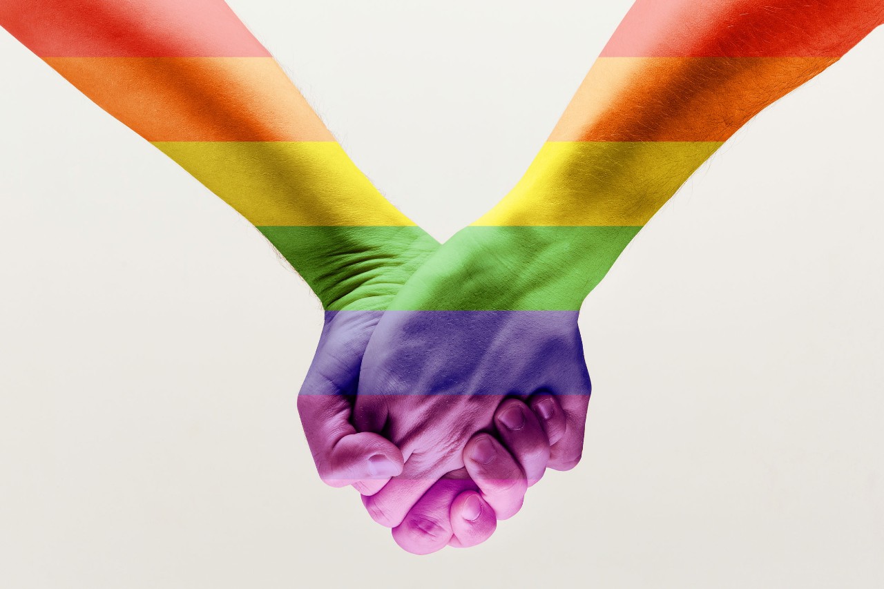 closeup of two clasped hands with overlay of rainbow flag