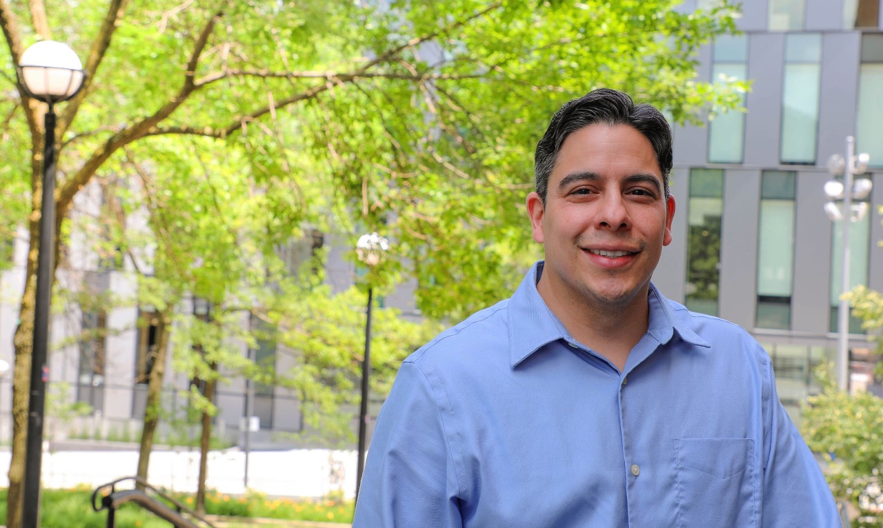 Man in blue dress shirt smiles and leans on wall with new Lindner Hall in background