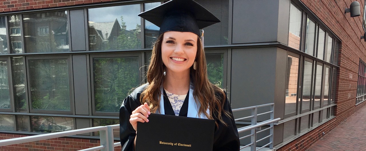 Amanda Bright, UC grad holds her graduation diploma on campus wearing her cap and gown.