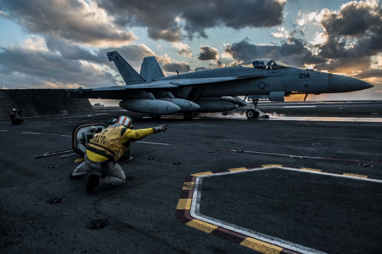 An F-18E Super Hornet takes off from the deck of the USS Ronald Reagan with the setting sun behind it.
