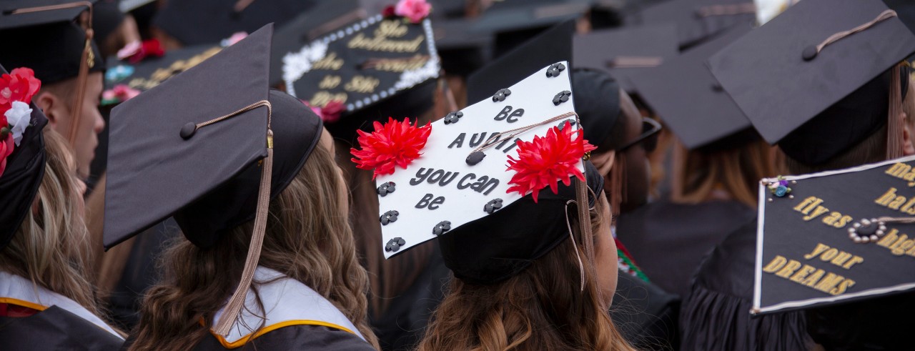 Image of UC graduates at Commencement wearing caps that have been decorated. One of them says "Be audit you can be." Another says "Fly as high as your dreams."