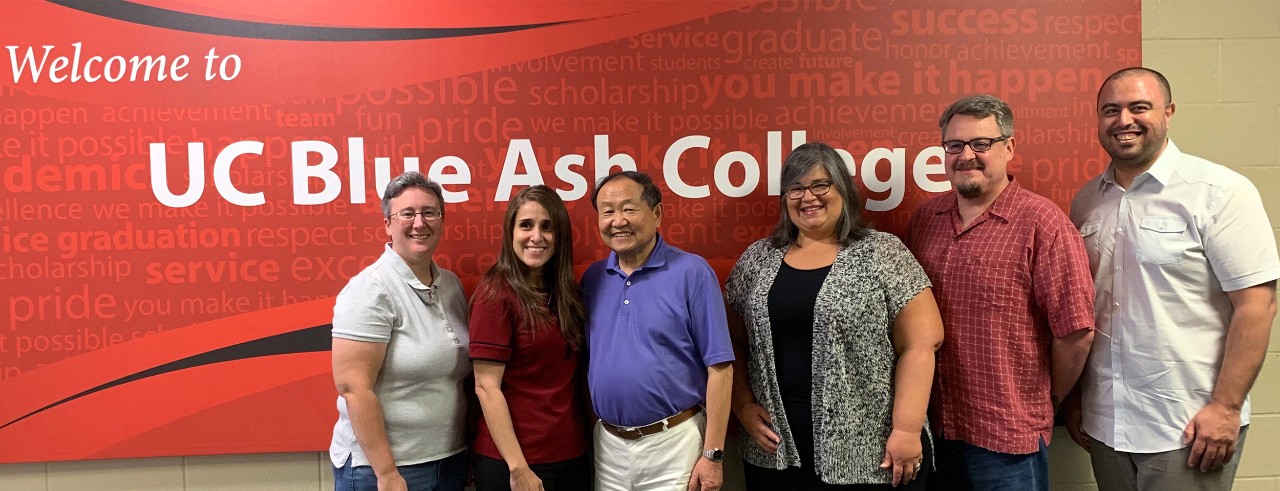 Six men and women stand in front of a sign reading, “Welcome to Blue Ash College.”