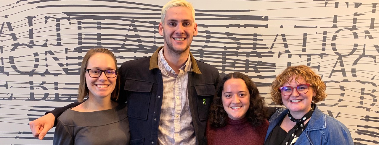 Four young people stand in front of a mural with unclear text