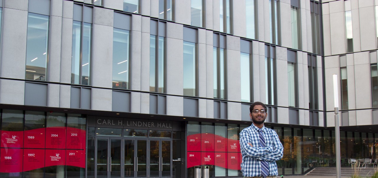 a man in a blue and shit checkered shirt and patterned blue tie stands in front of Carl H. Lindner Hall