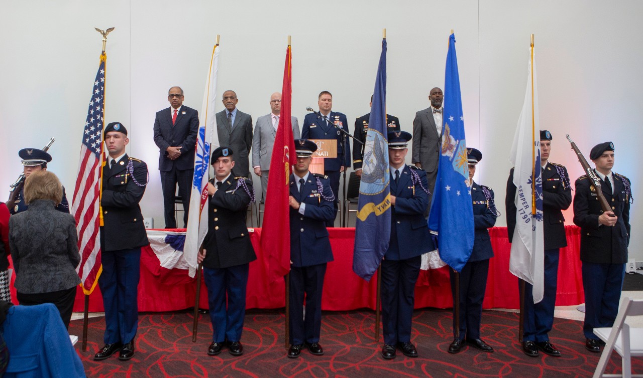 A military color guard standing in front of a stage, upon which several men in suits and two in military apparel also stand