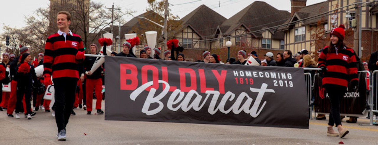 Students lead a parade holding a banner that says "Boldly Bearcat"