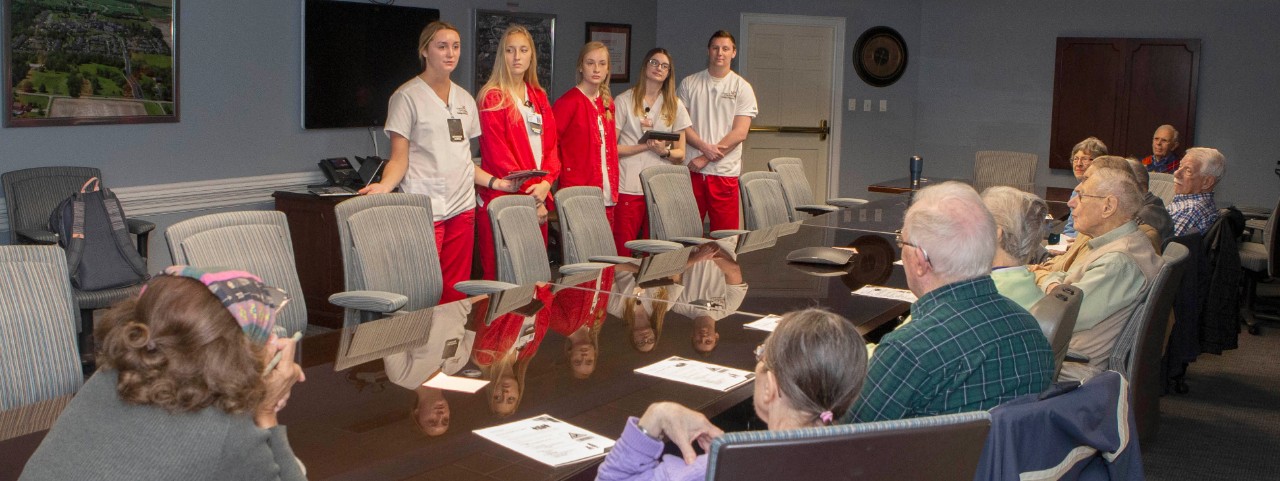 Five nursing students standing at a table across from several residents of Maple Knoll Village