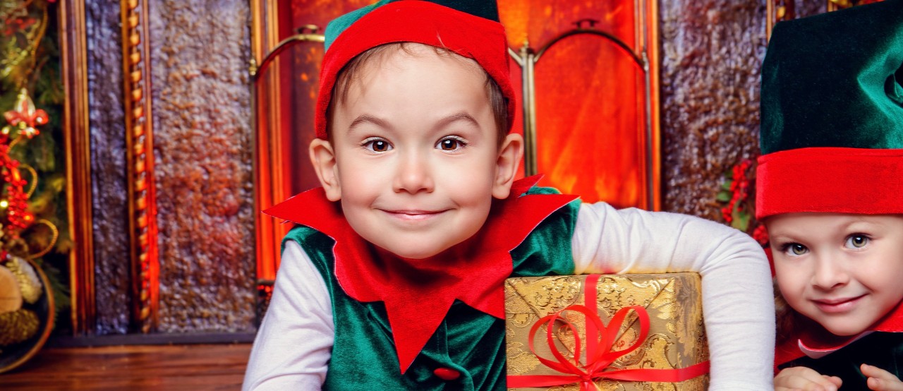 Two children dressed as Christmas elves smile while holding a wrapped present