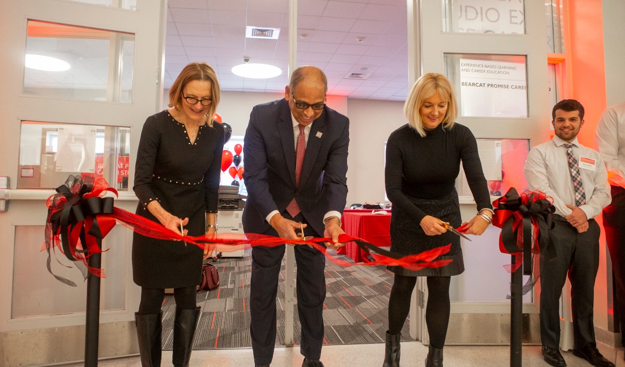 Two women in black dresses and one man in a suit cut a red and black ribbon with scissors