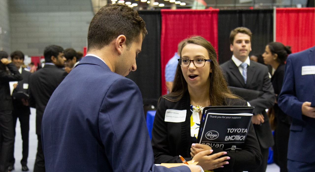 a woman with long dark hair and glasses and a suit jacket holds a binder and career fair packet while talking to a man in a blue suit jacket in an exhibition hall