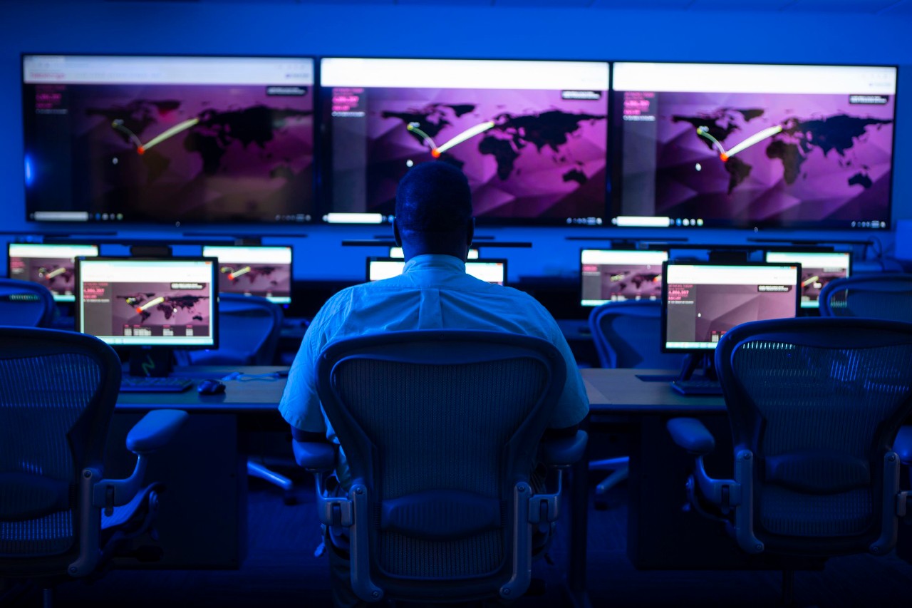 A man sits in an office chair in a dark room lit by computer screens