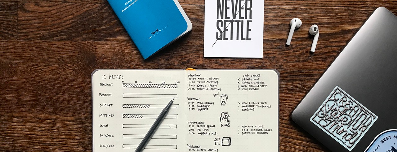 Two notebooks, airpods, a sticket and a laptop on a table. Sticker says "never settle."