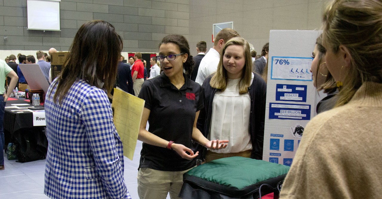 Four young women on a student team present an elevator pitch for a product to a judge in a science-fair-like setting in a gymnasium