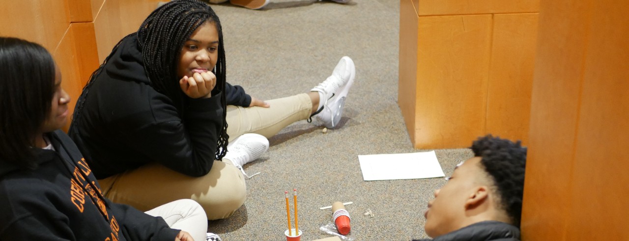 Three middle school students sit on the floor, building with pencils, plastic cups and cardboard.