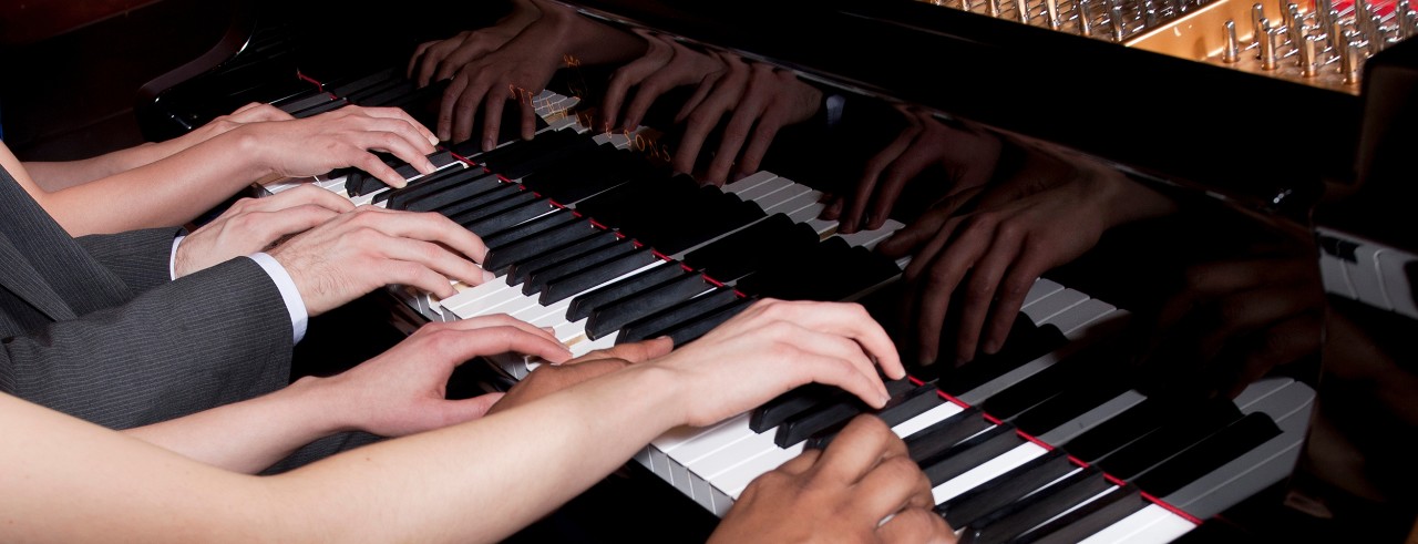 A close shot of multiple hands on a piano keyboard. 
