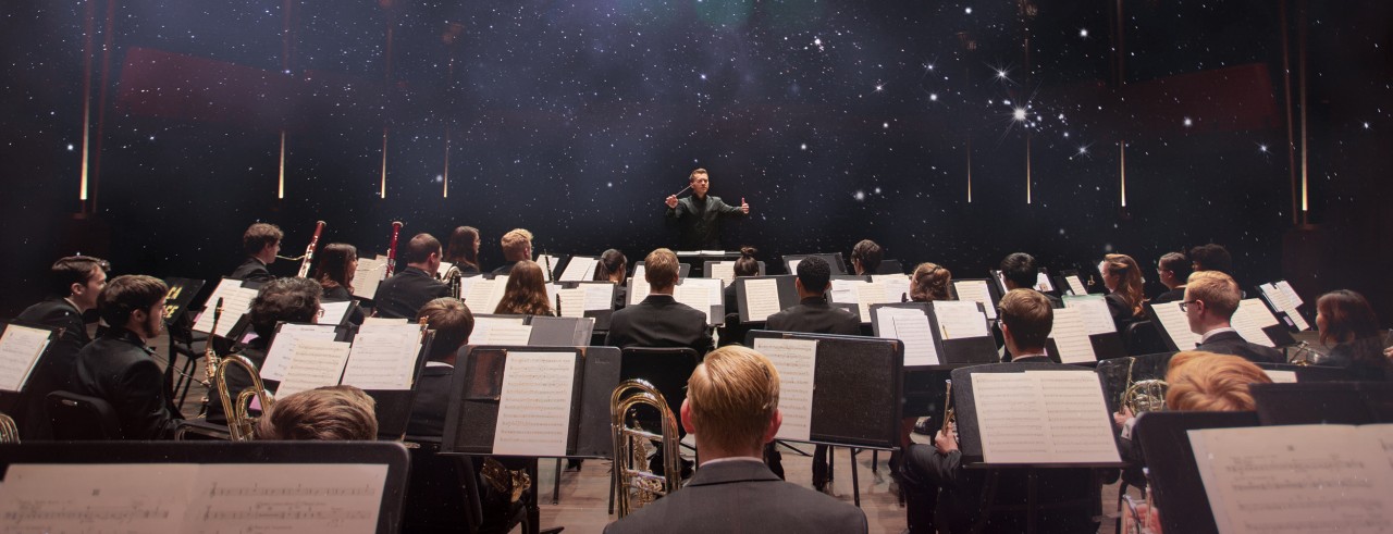 A photo of CCM Professor Kevin Michael Holzman conducting the CCM Wind Symphony as a starscape appears overhead.