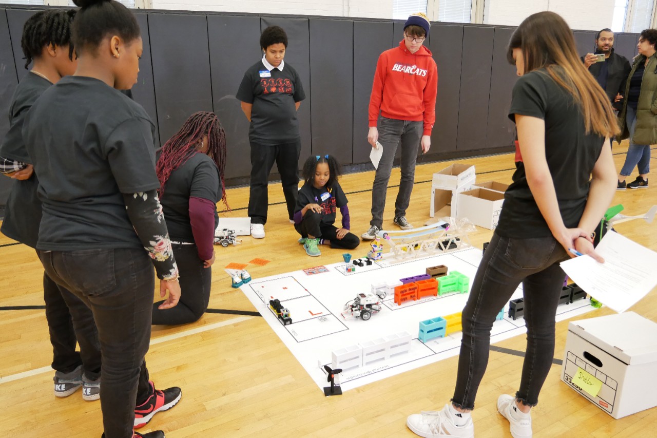 five elementary students and two judges watch a Lego robot navigate a course on the gym floor