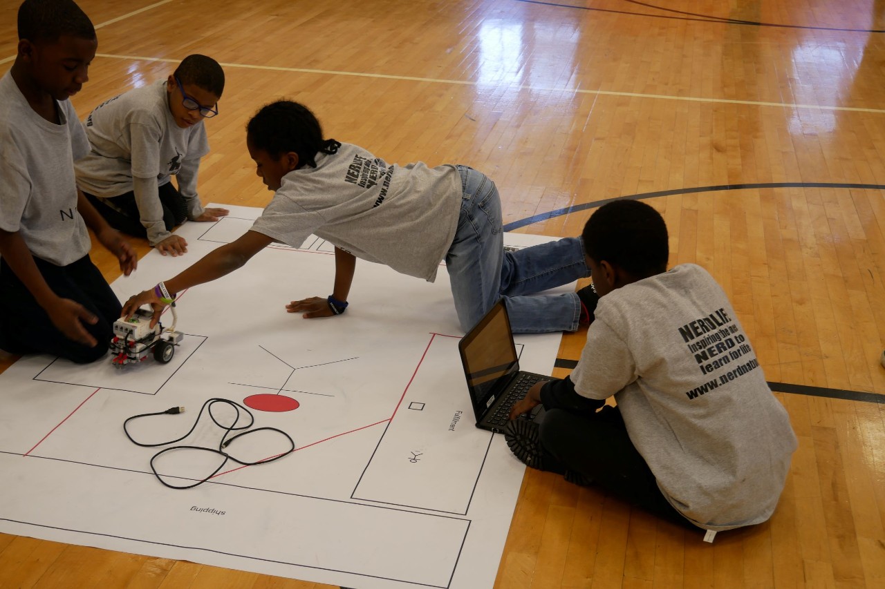 four elementary school students sit on the floor around a Lego robot, on a large paper floor plan 