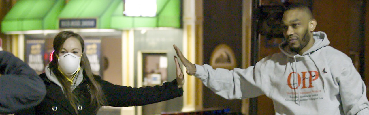 A woman wearing a black jacket and a medical mask reaches out to give a high five from a distance to a man wearing a grey sweatshirt and jogging pants