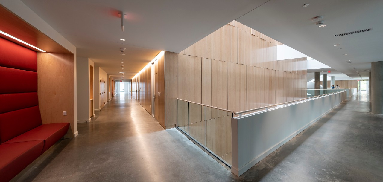 corner of two hallways inside Carl H. Lindner Hall. Red cloth booth off to left side. Pointed corner straight ahead with window well of light. Cement floors with white ceiling and light-colored wood