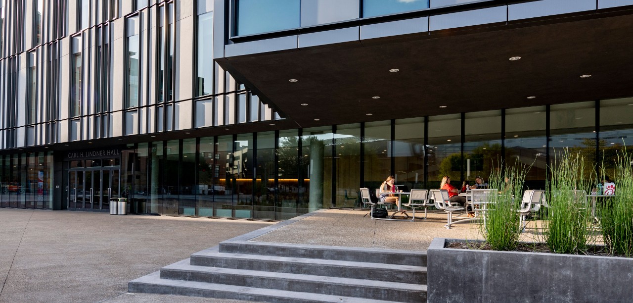 Two students sit working at a table outside the south entrance of Carl H. Lindner Hall in late afternoon/early evening. Green outdoor plants and steps are in the foreground