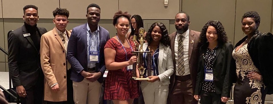 eight college students in business wear stand in a conference space, two in the middle hold a trophyding 