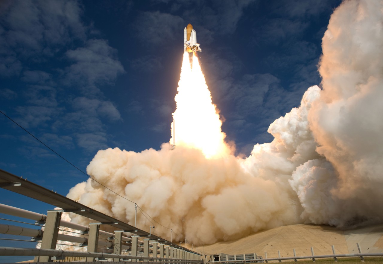 a photo looking up at a rocket being launched into the air, flames streaking under it and a cloud of exhaust blowing out behind it