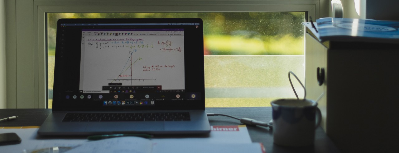 Laptop on desk at home in front of window with book and coffee cup.