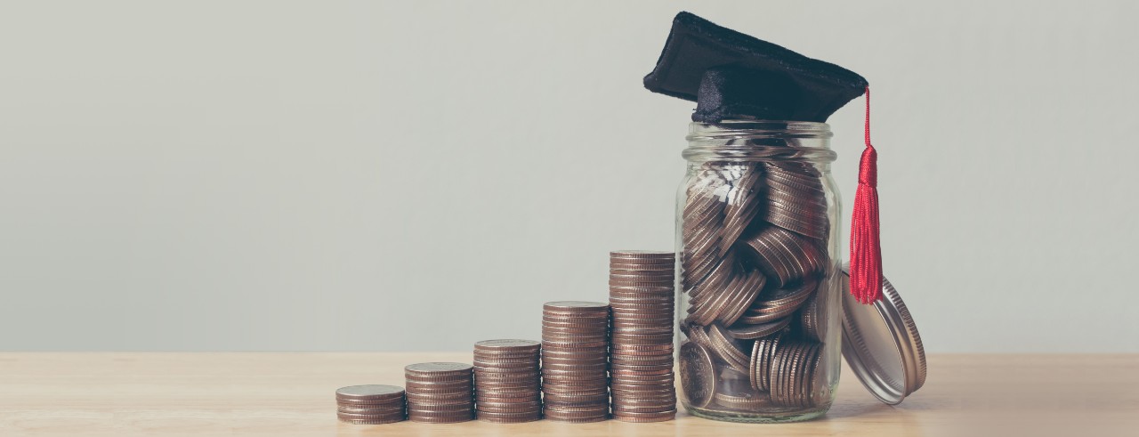 A miniature graduation cap sits atop a jar of quarters, which is next to a stair step of more quarters