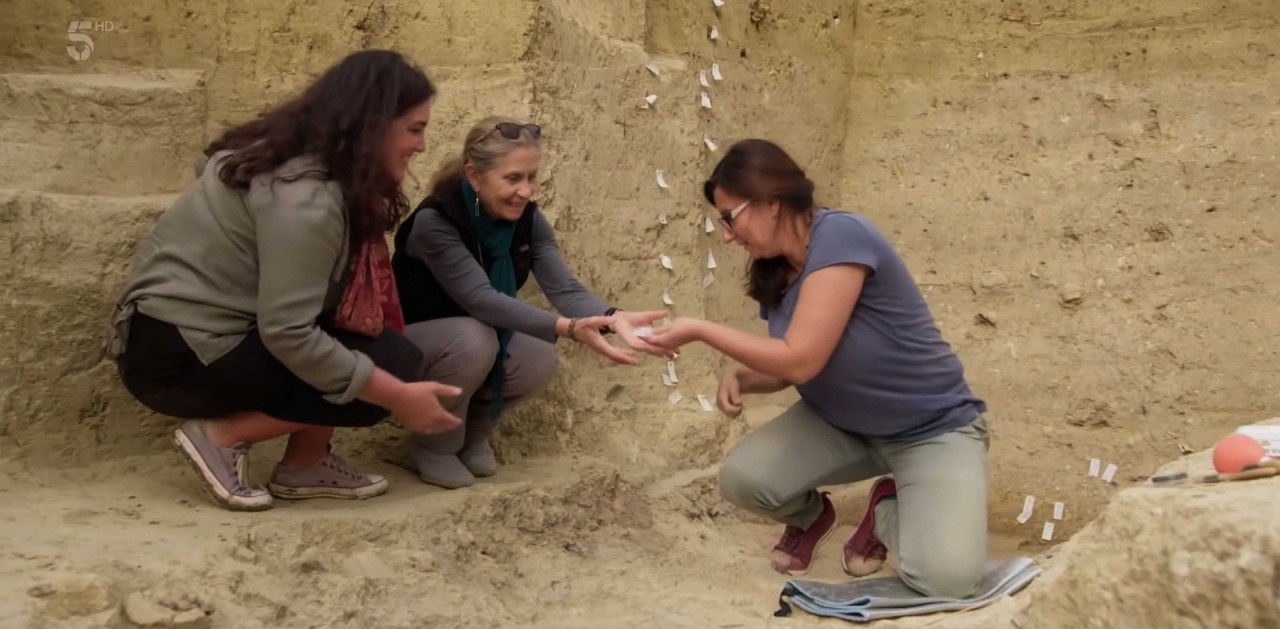 Channel 5 host Bettany Hughes, UC senior research associate Sharon Stocker and a Greek archaeologist kneel in an exposed tomb examining an artifact.
