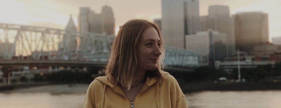 Erin Tepe leans against a railing and looks to her left while posing for a photo in front of the scenic Cincinnati skyline