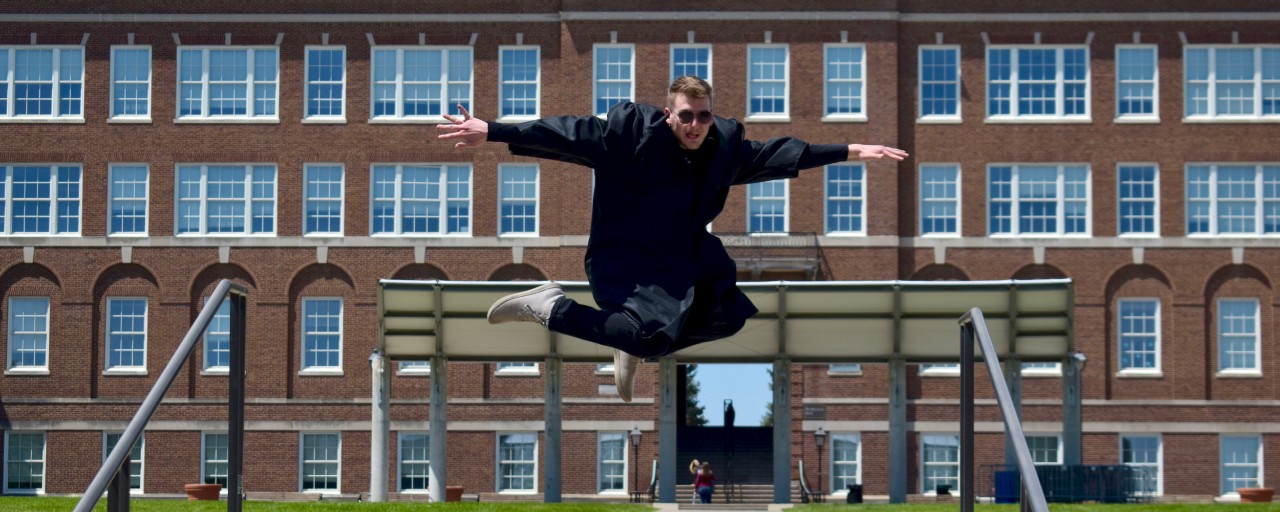 UC grad Ben Paulus jumps in the air wearing his graduation gown in front of McMicken Hall.