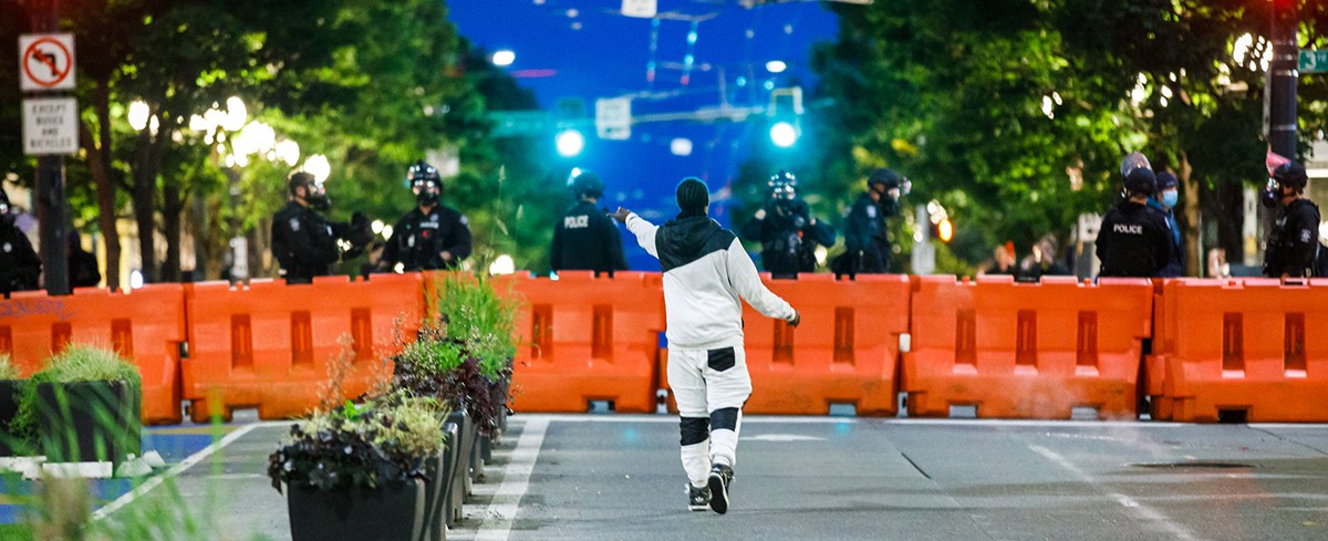 a protester stands in front of a line of police standing behind orange barricades